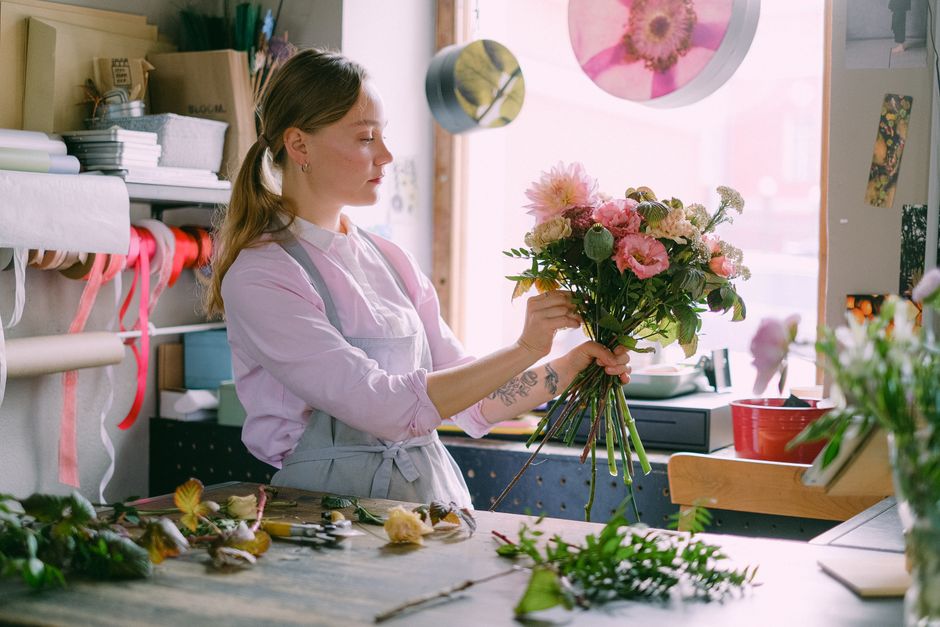 A person with dark hair, wearing a black sweater, is arranging a large bouquet of fresh flowers on a white-covered table. The bouquet features a mix of pink and white blooms, including snapdragons and