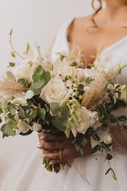A close-up of a fresh floral bouquet held by a person wearing a white long-sleeve shirt. The arrangement features soft pink and white flowers, including azaleas or similar blooms, complemented by dark