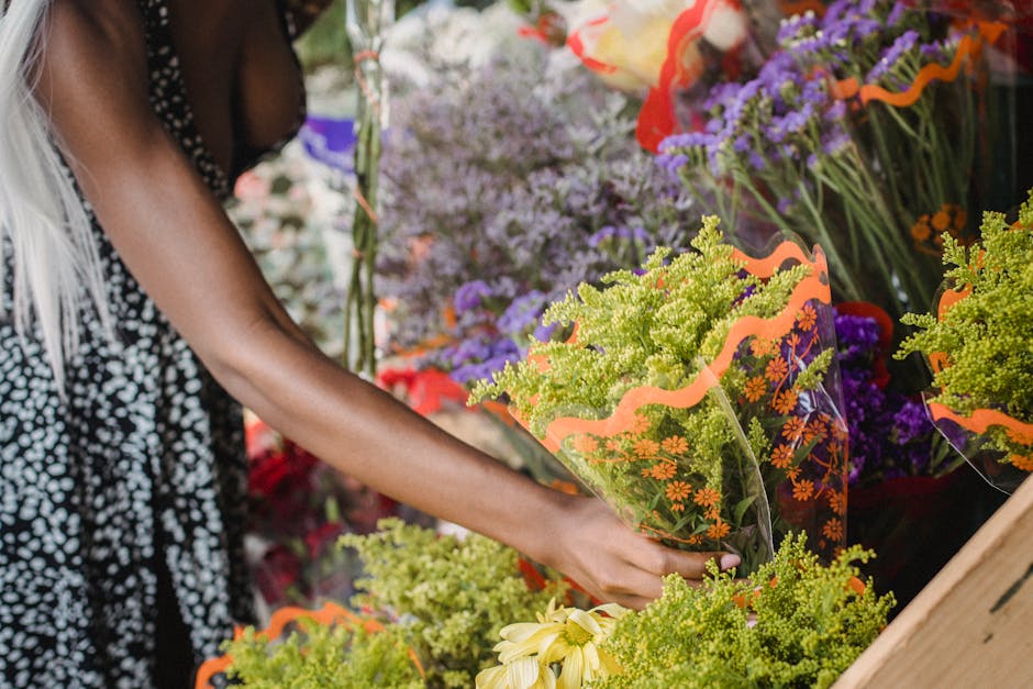 A woman with braided, silver-gray hair wearing a maroon fleece jacket and bright blue gloves is closely inspecting a bouquet of white flowers wrapped in clear plastic. She is in an outdoor flower mark