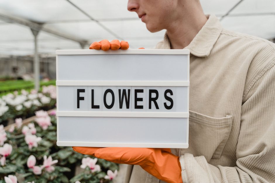 A person wearing an off-white shirt and orange gloves is holding a white lightbox with the word 'FLOWERS' displayed in black letters. The individual is standing in a greenhouse surrounded by rows of b