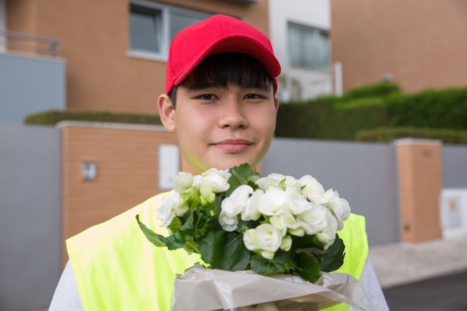 A woman with long dark hair, dressed in a vibrant red top, smiling as she receives a bouquet of fresh white flowers wrapped in white paper from a delivery person wearing a high-visibility vest and red
