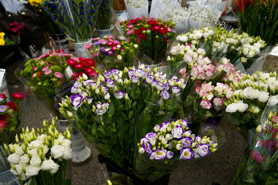 A variety of fresh flower bouquets arranged on a display table, featuring pink, purple, white, and red blossoms including lisianthus, roses, and small daisies. The flowers are wrapped in clear celloph